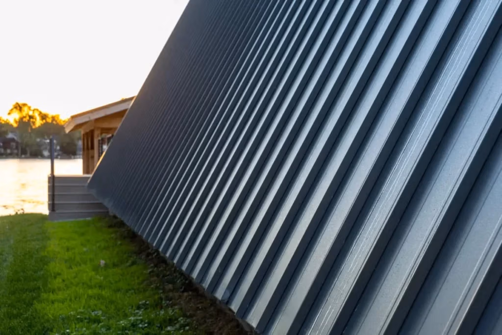 Close-up of dark gray corrugated standing seam metal siding or roofing panels angled diagonally over green grass, with a wooden structure and golden sunset reflecting on a lake visible in the background.