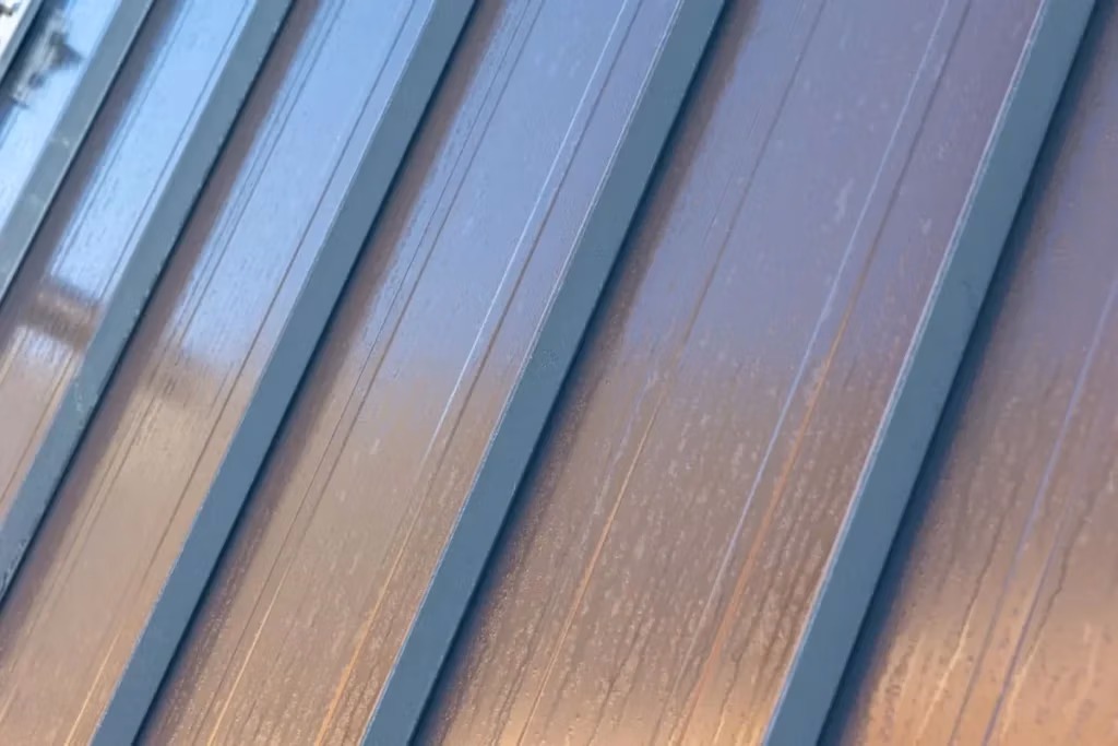 Close-up view of dark charcoal blue standing seam metal roofing panels with raised seams and a warm brown oxidized sheen reflecting sunlight.