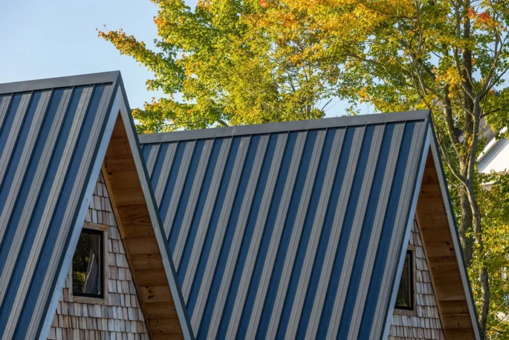 Two steep A-frame cabin rooftops clad in blue corrugated standing seam metal panels, flanked by cedar shake siding and small gable windows, set against autumn foliage trees and a clear sky.