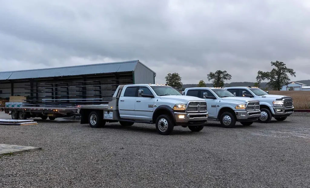Three white Ram heavy-duty pickup trucks parked on a gravel lot in front of a metal building, with the lead truck hauling a flatbed trailer loaded with steel roofing panels on an overcast day.