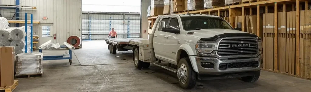 A white RAM 5500 heavy-duty flatbed truck pulls a loaded trailer through the interior of a large industrial warehouse stocked with lumber and building materials, with a worker visible in the background near a loading dock.