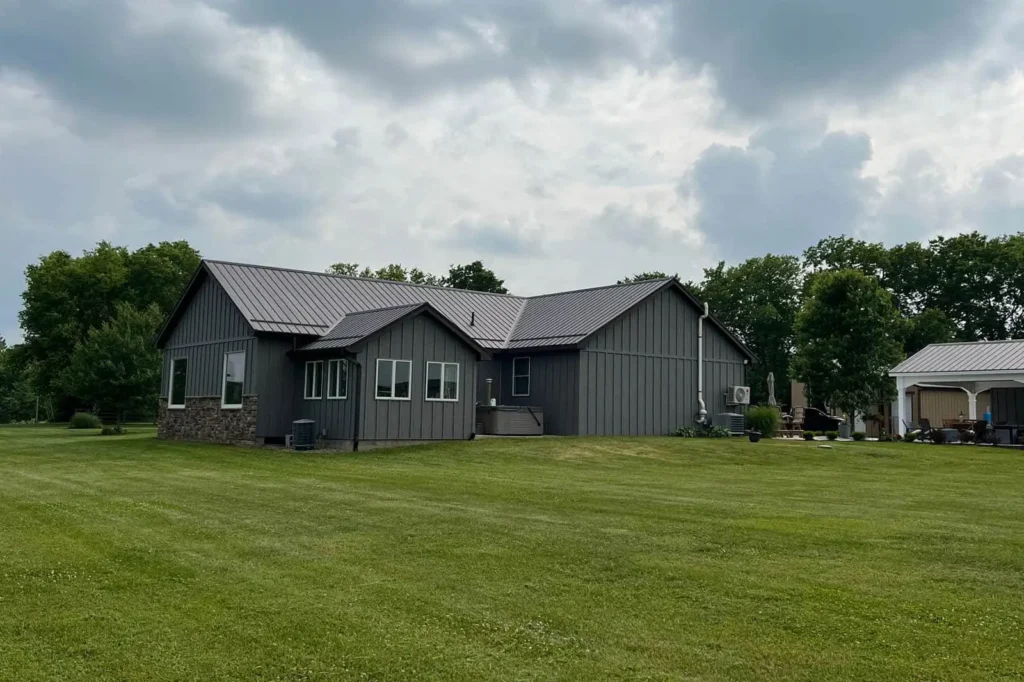 A modern single-story home with dark gray board-and-batten vertical siding and a matching dark metal standing-seam roof, surrounded by a well-manicured green lawn with mature trees in the background under a cloudy sky.