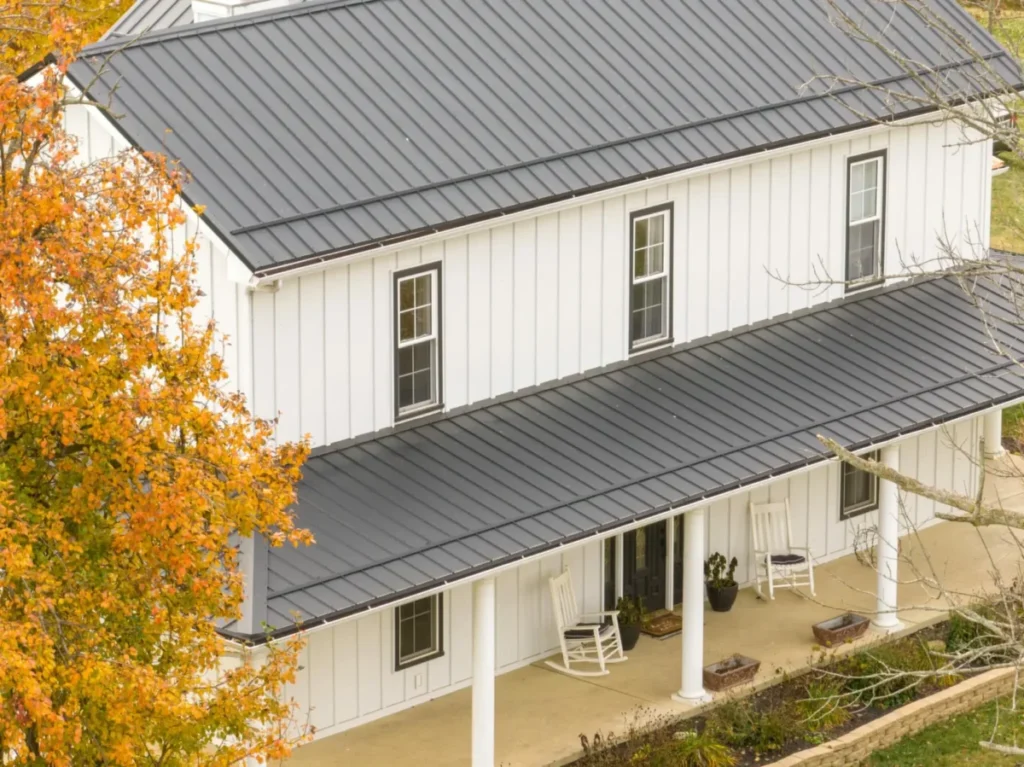 Aerial view of a two-story white board-and-batten farmhouse with a dark gray standing seam metal roof and a covered front porch with white rocking chairs, surrounded by autumn foliage.