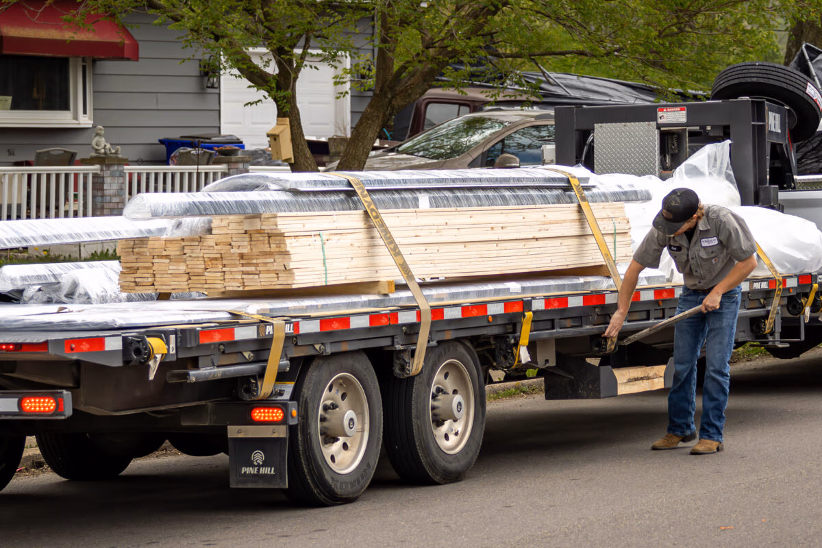 Flatbed truck loaded with stacked lumber and plywood, with worker standing beside it on residential street