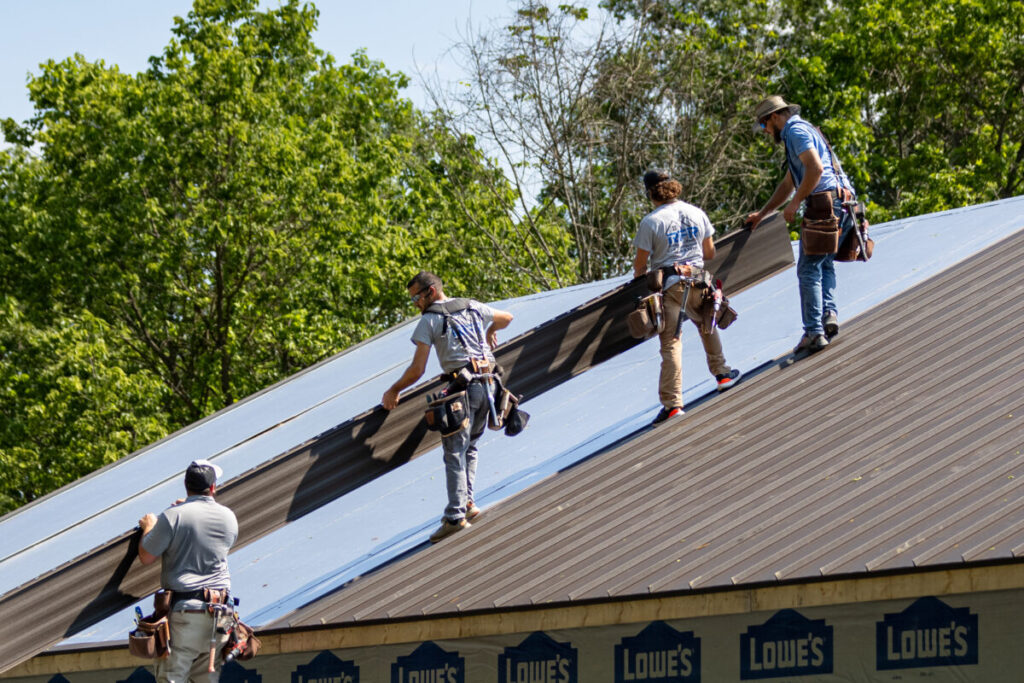 Four workers in tool belts carry and position large dark metal roofing panels on a steep residential or commercial roof, with Lowe's-branded materials visible along the eave and green trees in the background.