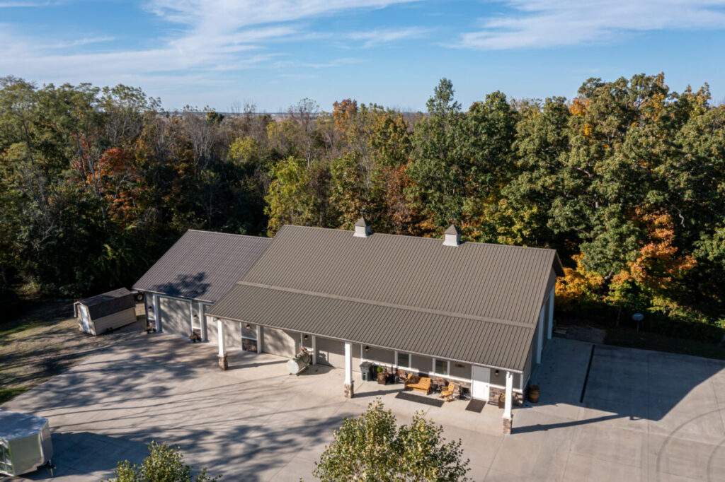 Drone aerial photograph of a large single-story barndominium with a dark gray standing-seam metal roof, covered front porch with white columns, and multiple garage bays, surrounded by autumn-colored trees and a wide concrete driveway.