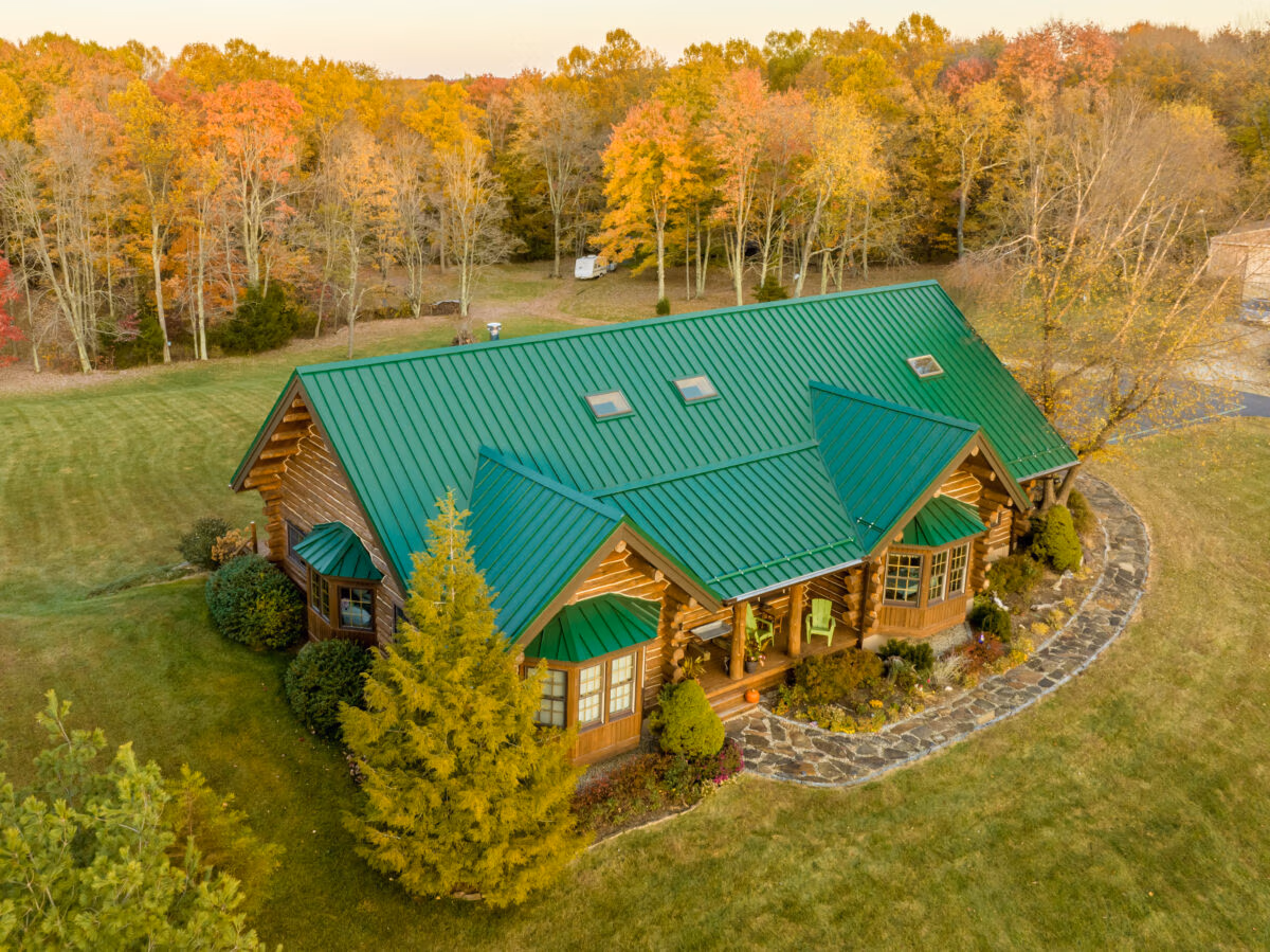 Aerial view of a large residential home with green standing seam metal roof and matching siding, surrounded by deciduous trees in fall foliage