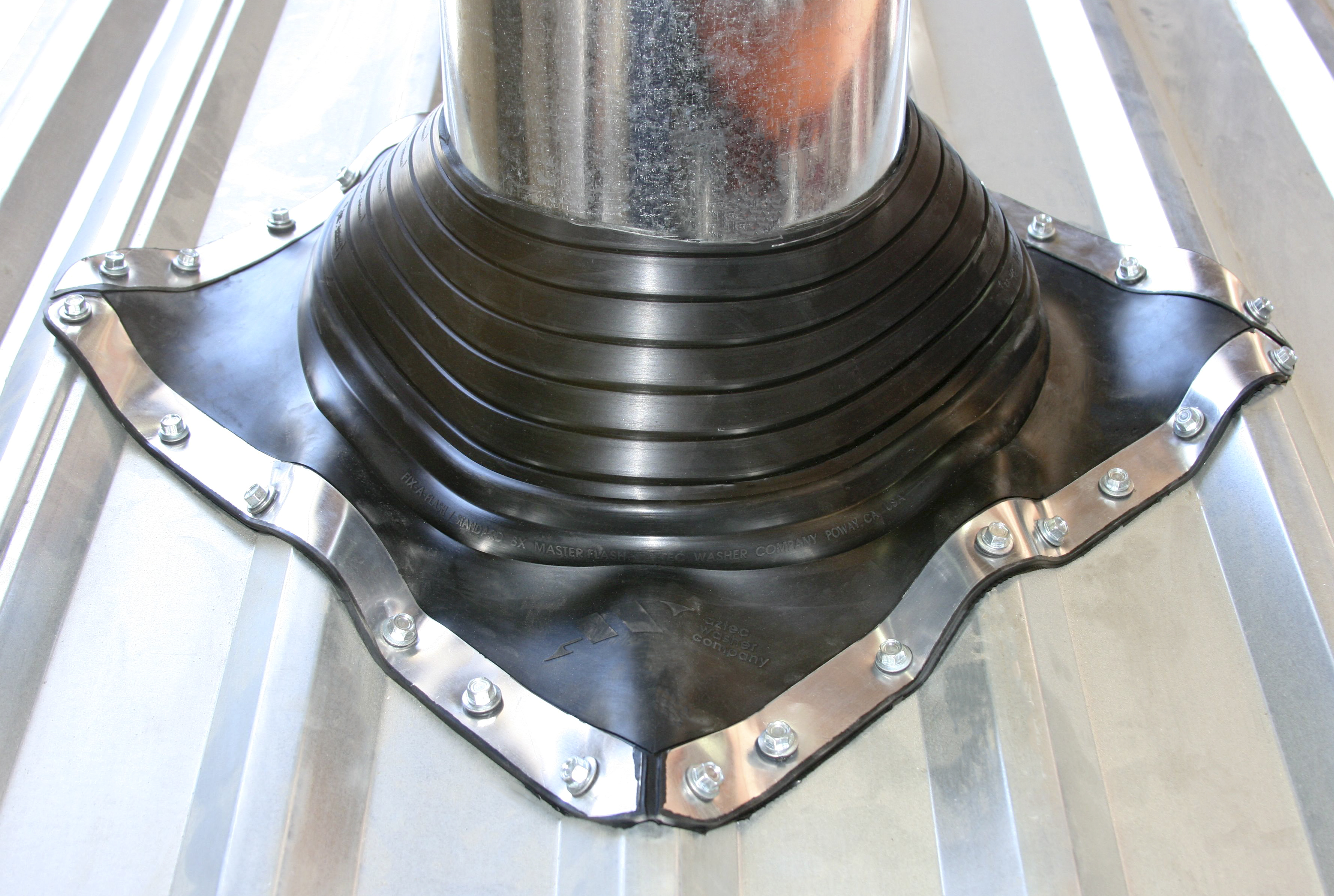 A black rubber pipe boot flashing seal installed around a metal chimney or vent pipe on a corrugated metal roof, secured with screws and a stainless steel retaining band around its perimeter.