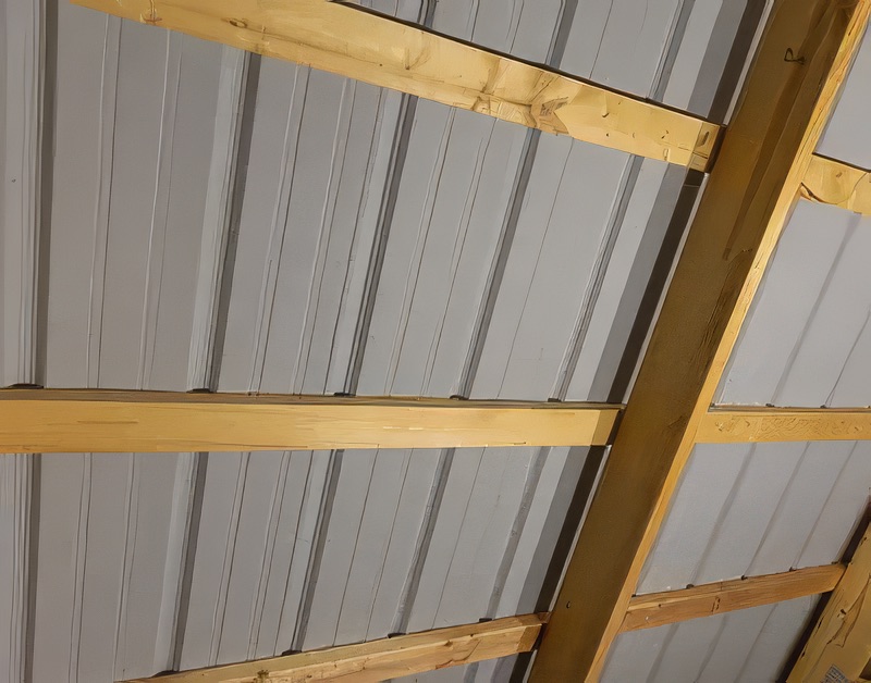 Interior view looking up at a corrugated gray metal roof with evenly spaced horizontal yellow pine wood purlins or girts spanning across the rafters of a pole barn or agricultural building.