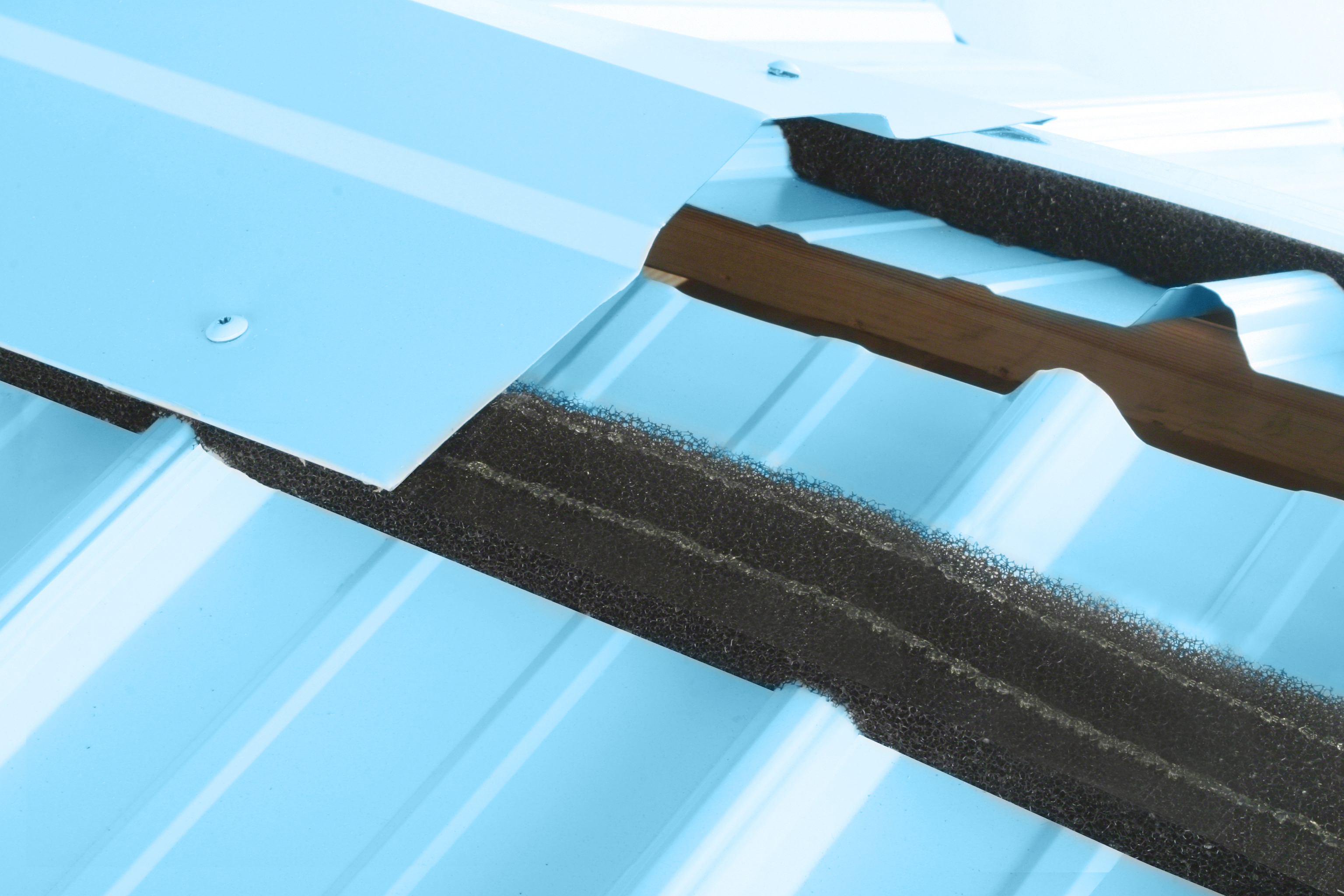 Close-up of black foam closure strips being installed between corrugated light blue metal roofing panels at the ridge cap area.