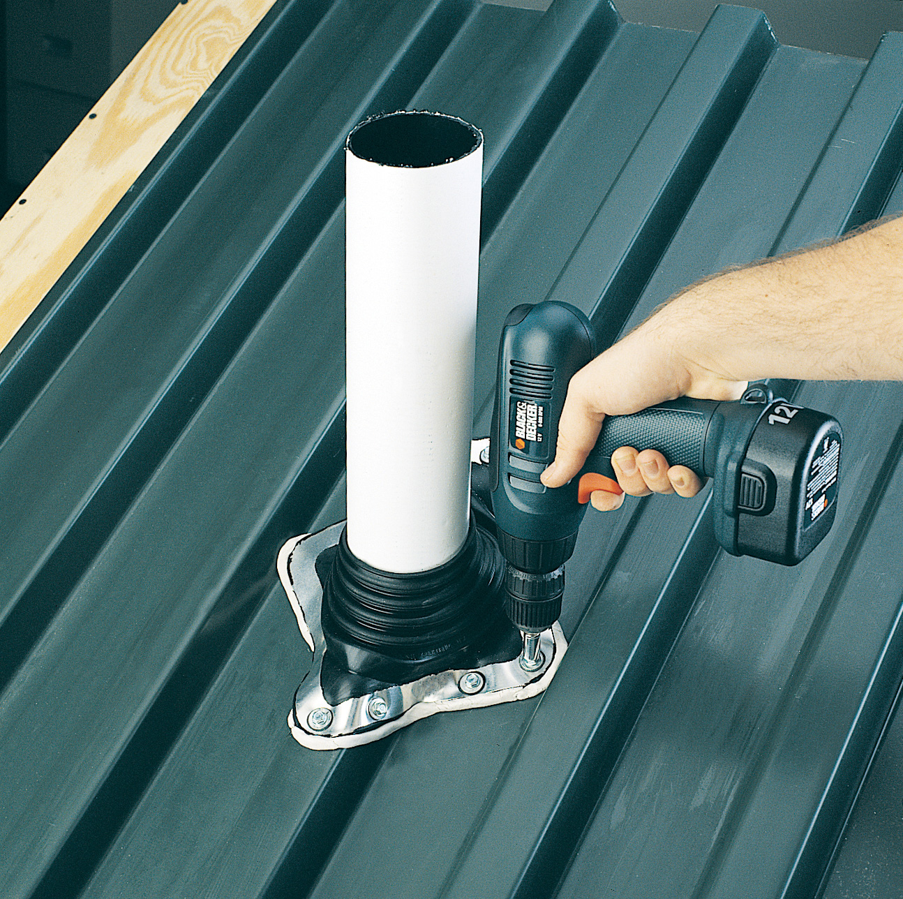A hand holding a cordless drill fastens a white PVC pipe through a rubber roof jack flashing boot mounted on a dark green corrugated metal roof.