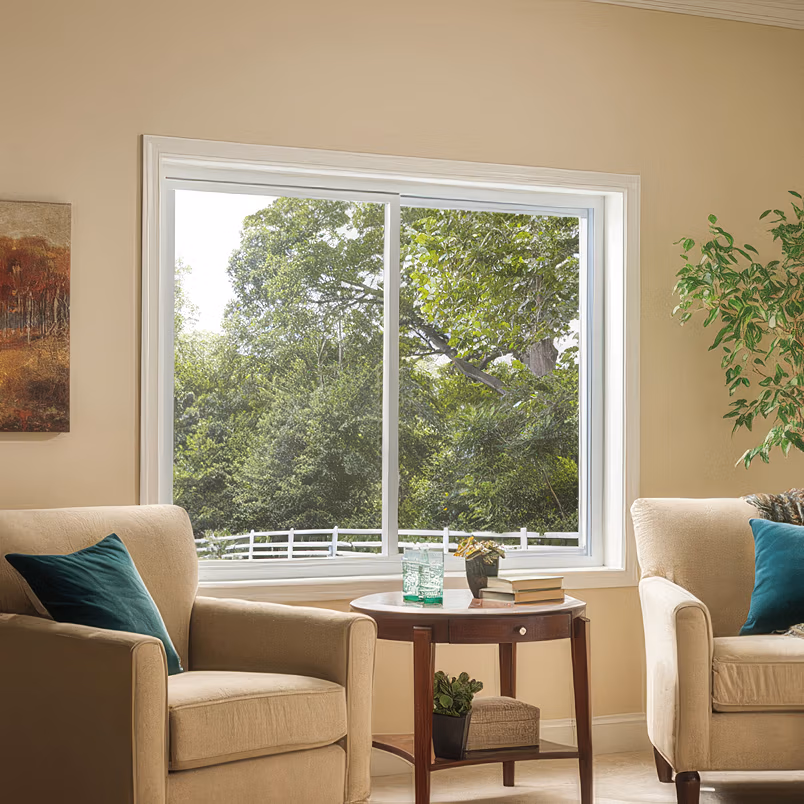 Bright living room with two cream-colored armchairs flanking a side table, viewed through large multi-pane gliding windows with white frames overlooking green trees.