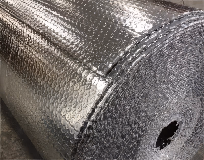 A large roll of silver reflective bubble wrap insulation with a dimpled metallic foil surface, shown on a concrete floor.