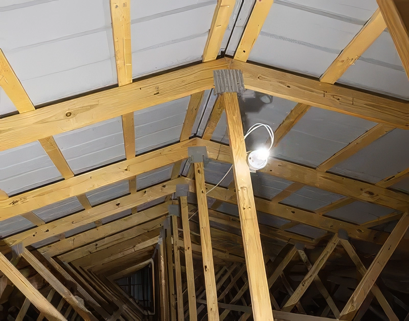 Interior view looking up at wooden roof trusses with white spray-applied moisture barrier or underlayment visible on the roof deck, illuminated by a single hanging work light in an unfinished building.