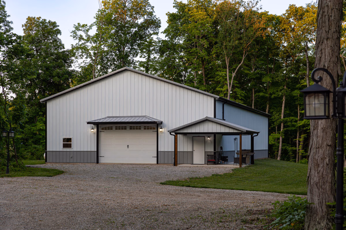 Modern pole barn with metal siding and metal roof, single-car attached garage, surrounded by trees and gravel driveway