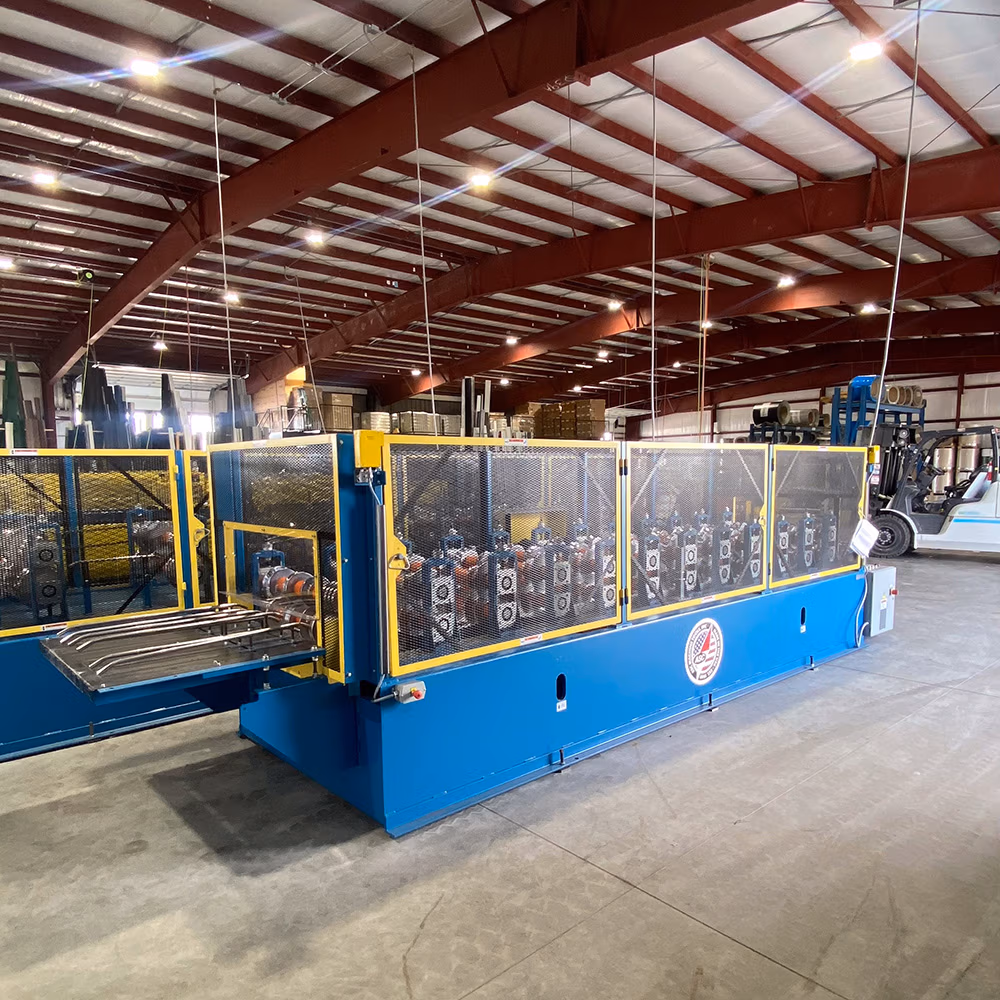A large blue and yellow roll forming machine with safety mesh guards sits on a concrete floor inside a steel-framed industrial warehouse with red beams and overhead LED lighting.