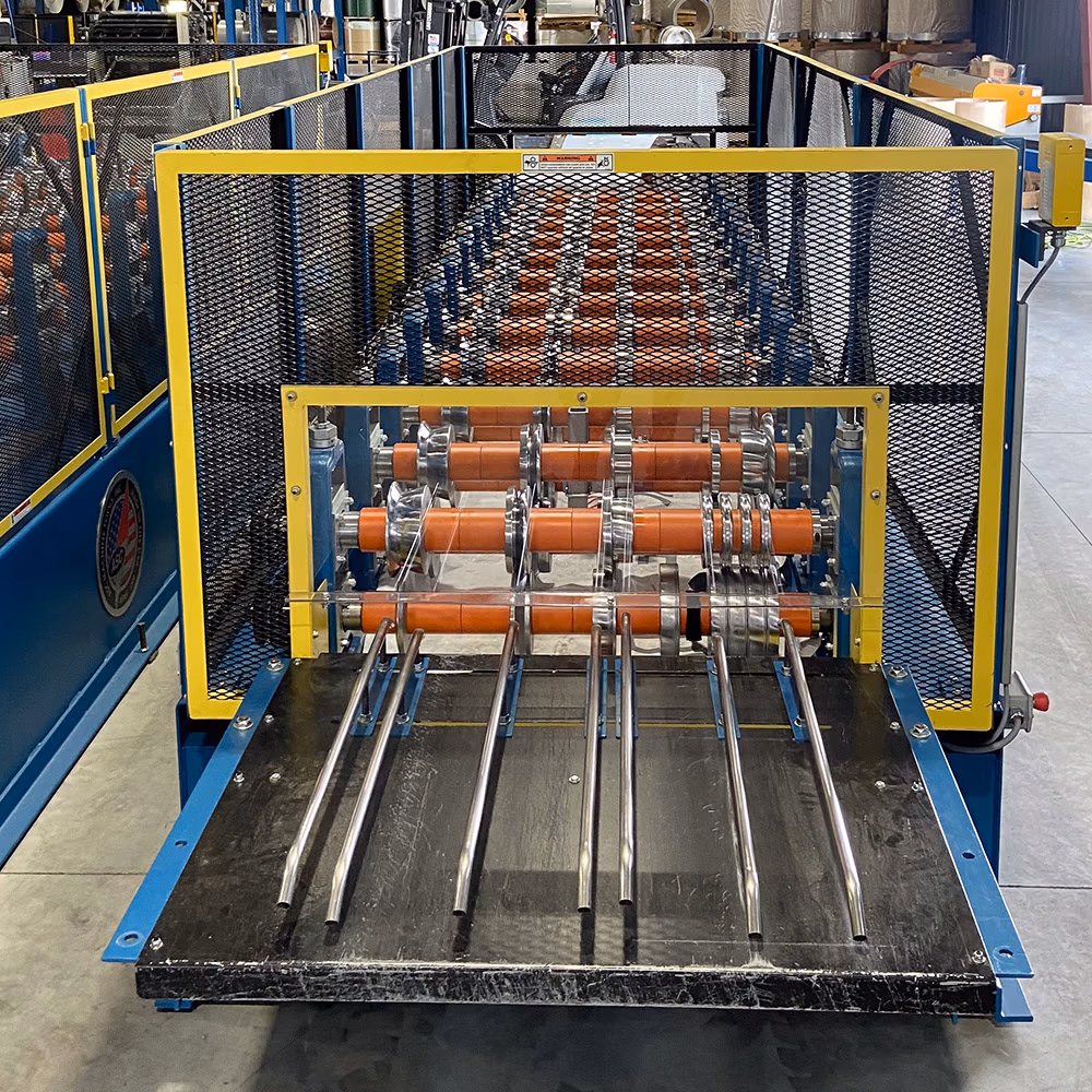 A blue and yellow industrial roll forming machine with orange polyurethane rollers, stainless steel guide rods, and yellow mesh safety guards in a manufacturing facility.