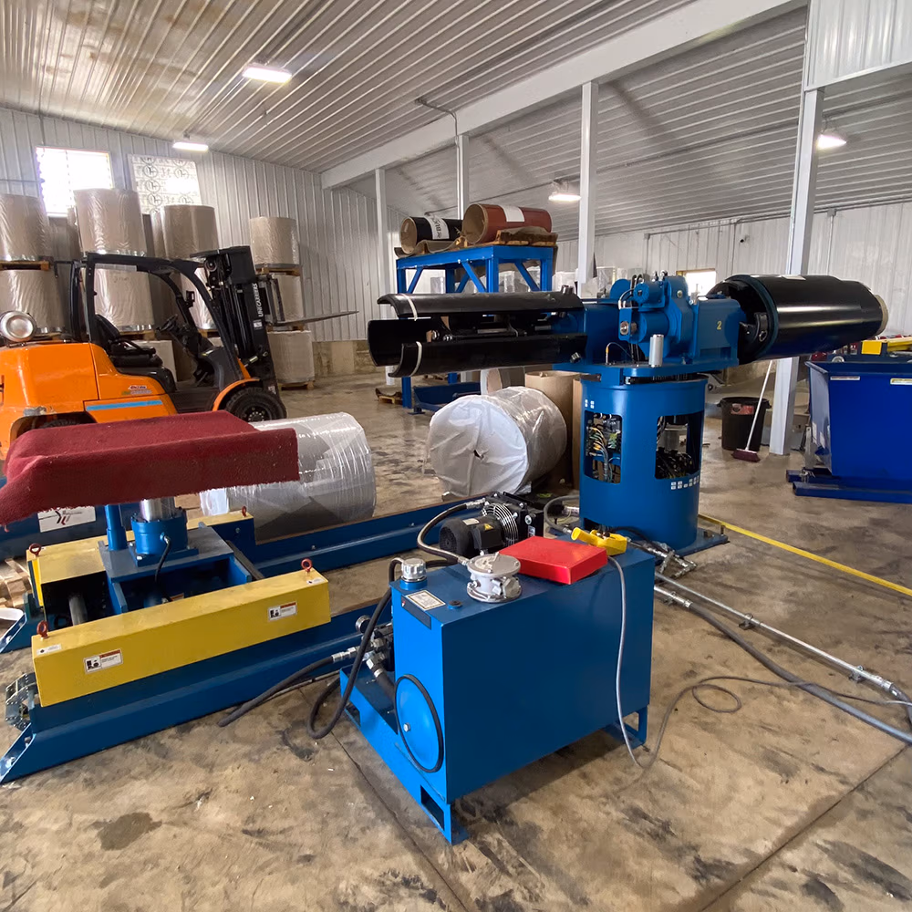 Blue hydraulic roll handling and unwinding machinery with yellow and red controls positioned on a concrete floor inside a metal-walled industrial warehouse, with an orange forklift visible in the background.