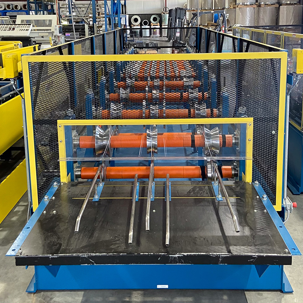 A blue and yellow industrial slitter rewinder machine featuring orange rubber rollers, metal separating guides, and a clear acrylic safety guard, photographed in a manufacturing facility.