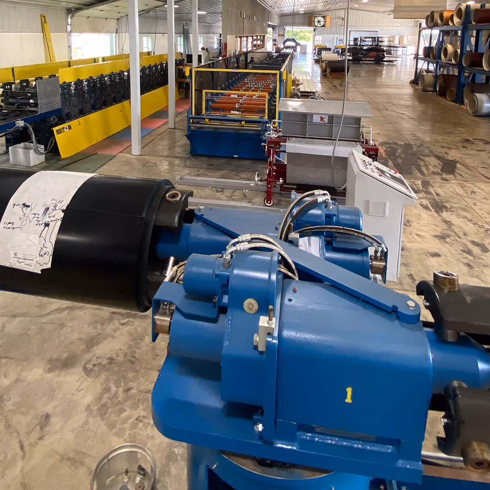 A large industrial manufacturing facility featuring a blue hydraulic decoiler holding a black coil of sheet metal in the foreground, with a roll forming production line stretching deep into a spacious warehouse.