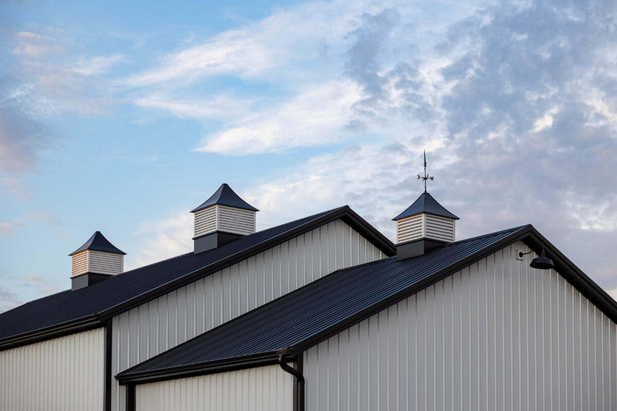 A modern white metal barn building with three decorative white cupolas topped with dark pyramid roofs against a blue cloudy sky, with one cupola featuring a weather vane.