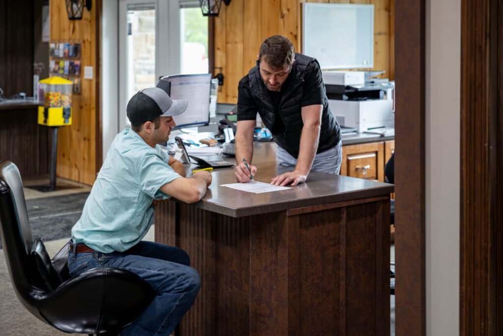 A man in a black vest leans over a dark wood reception counter to sign a document while a seated employee in a light blue shirt and gray snapback cap watches at a wood-paneled office interior.