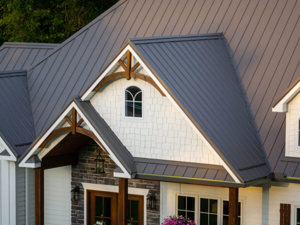 A craftsman-style farmhouse exterior featuring a gray standing seam metal roof with decorative wooden gabled brackets, white shake siding, stone veneer accents, and arched attic window.