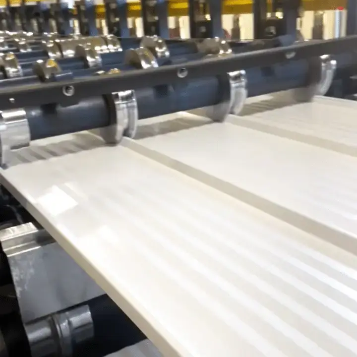 A white corrugated metal panel moves through a roll forming machine with chrome rollers and a dark blue steel frame in an industrial manufacturing facility.