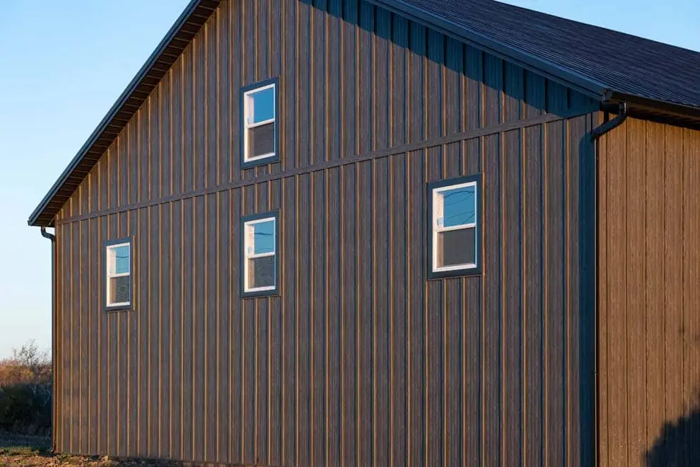A large barn-style building clad in dark brown wood grain vertical board and batten metal siding, featuring four white-trimmed double-hung windows on the side wall under a dark metal roof.