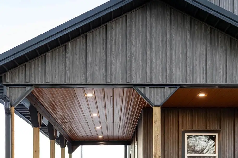 Close-up of a modern building exterior featuring gray wood grain Aristolath board and batten siding on a gabled facade, with a warm brown tongue-and-groove soffit ceiling illuminated by recessed lighting on a covered carport or porch area.
