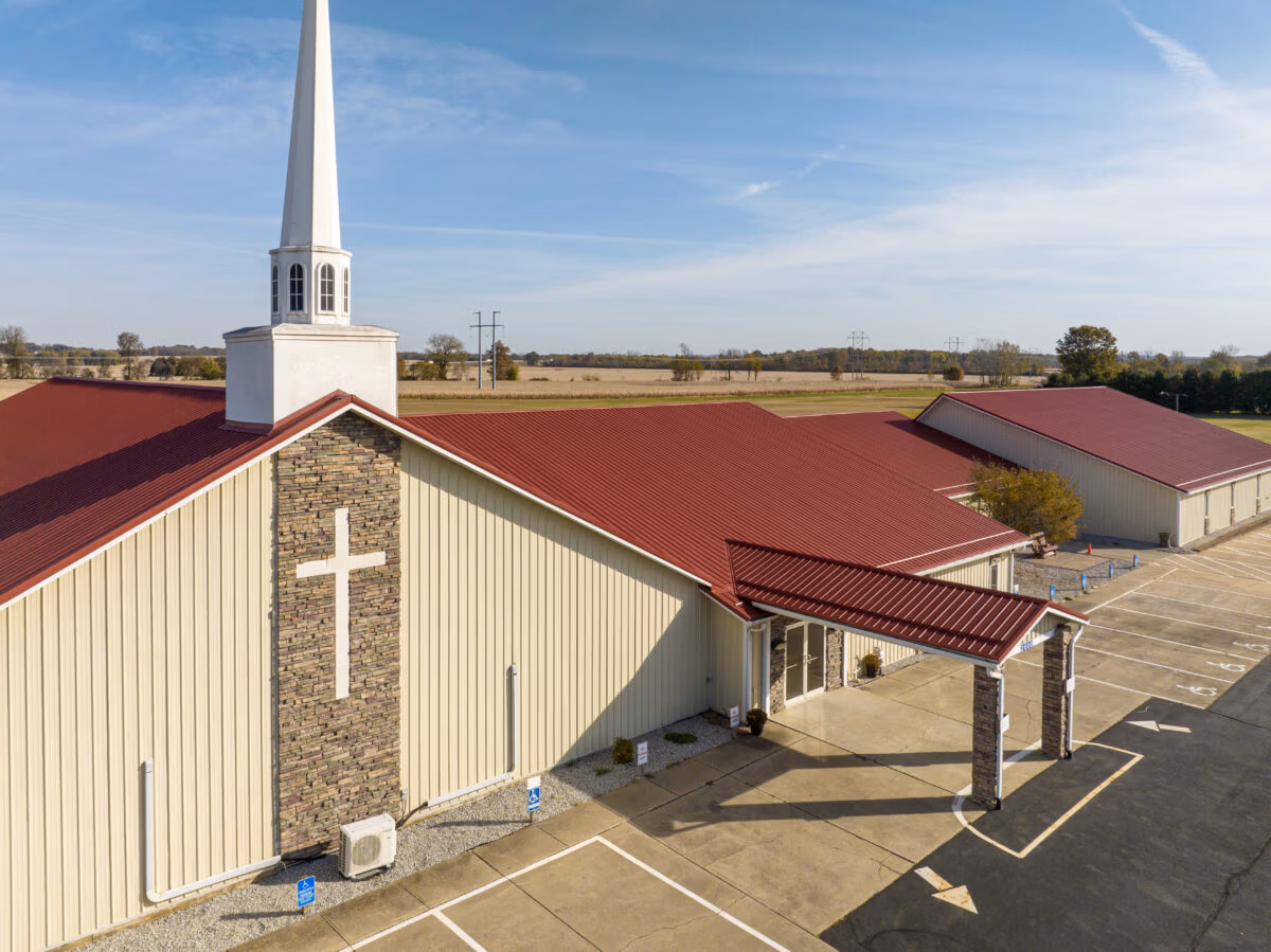Aerial drone view of a cream-colored metal sided Christian church with a white steeple, red standing-seam metal roof, stone accent walls, a white cross, covered entrance canopy, and an empty parking lot surrounded by flat agricultural farmland.