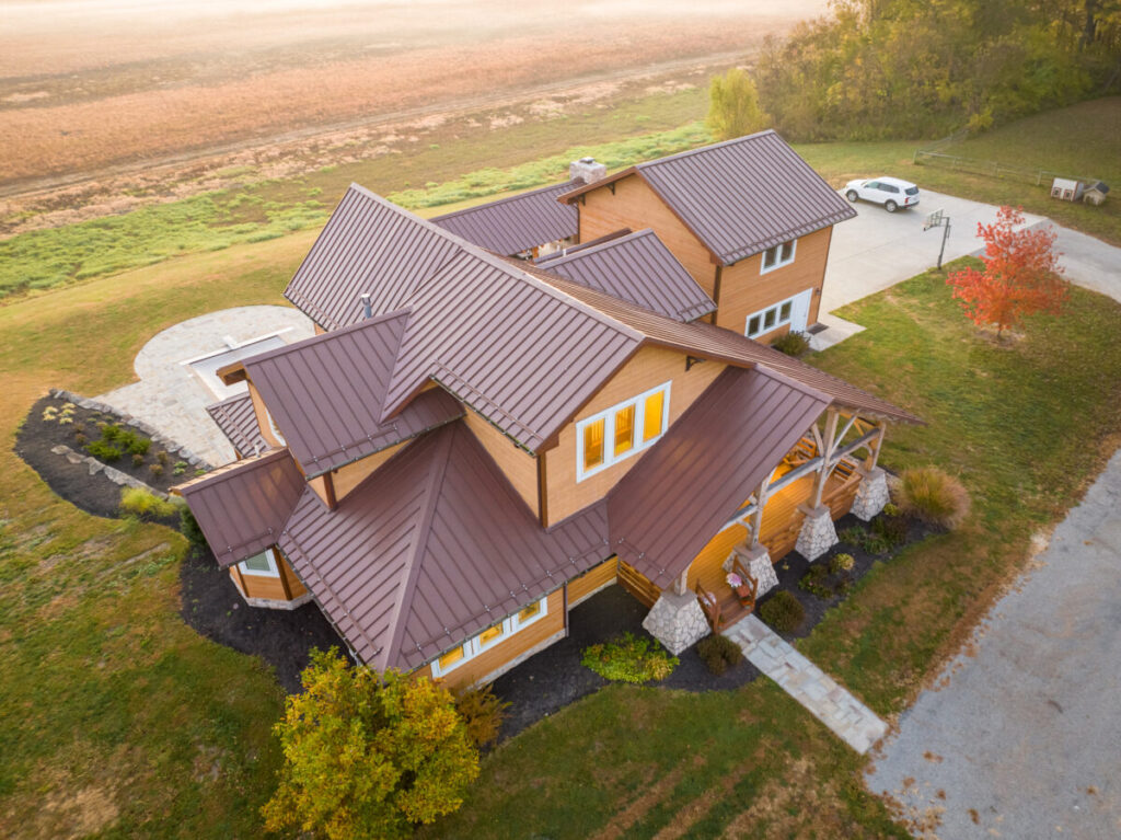 Aerial drone photograph of a large timber-frame house with a complex brown standing-seam metal roof, warm wood siding, stone porch columns, and illuminated windows, surrounded by green lawns and autumn trees in a rural setting.