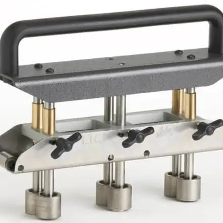 A Malco three-station edge roller tool with a black ergonomic handle, brass spring-loaded posts, stainless steel adjustable thumb screws, and cylindrical rolling dies mounted on a metal base plate.