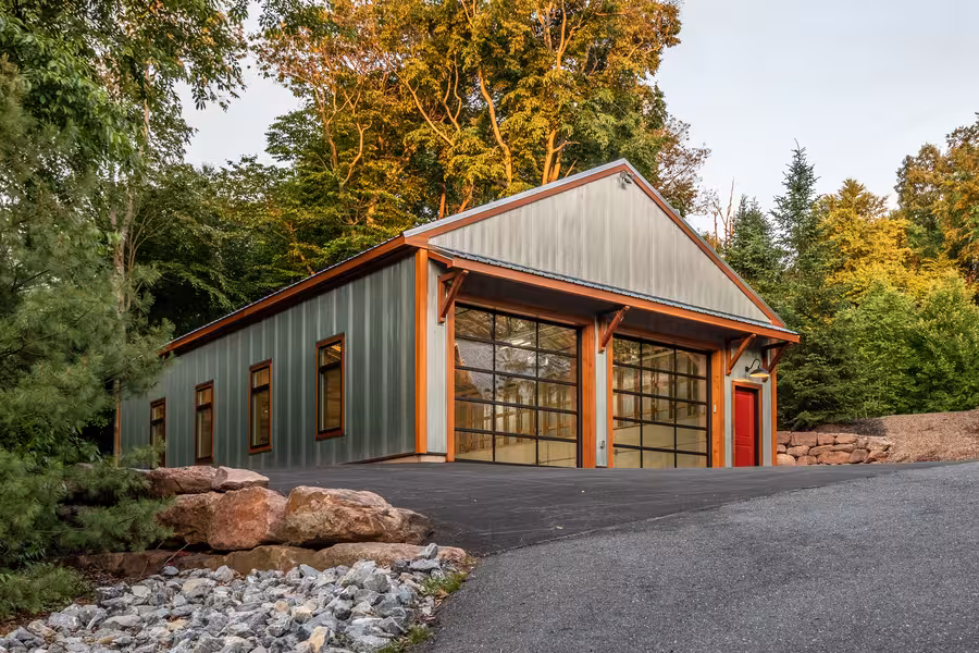Contemporary barn-style building with corrugated metal siding, standing seam roof, large industrial windows, and red entry door surrounded by trees.
