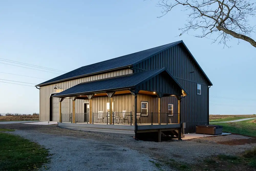 A large dark gray metal barndominium with a black standing seam roof, warm-lit covered front porch with wood beam columns and black railing, photographed at dusk on a rural gravel property.