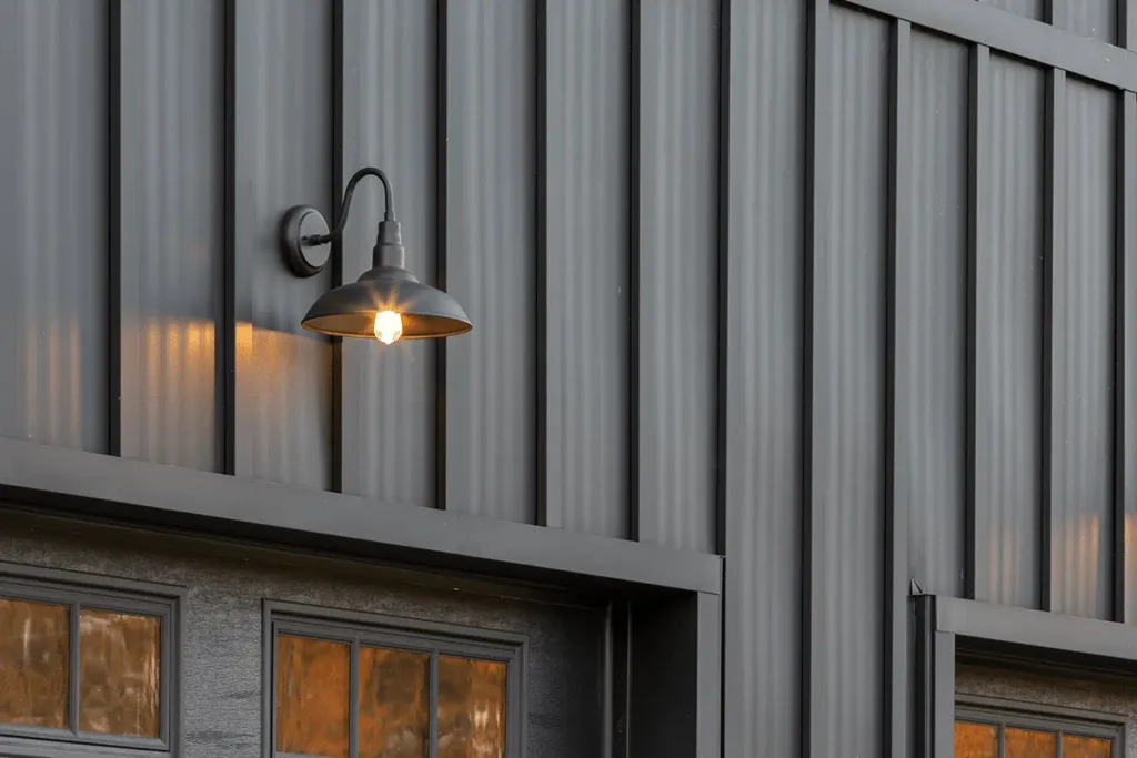 A black gooseneck barn wall sconce with an illuminated Edison bulb mounted on dark gray corrugated metal board and batten siding above garage doors with warm amber-lit windows.