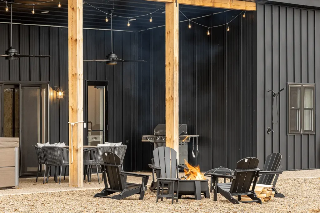 A modern black metal board-and-batten barndominium exterior features a covered patio with natural wood posts, string lights, black Adirondack chairs围 around a lit fire pit, a gas grill, and a gravel ground surface.