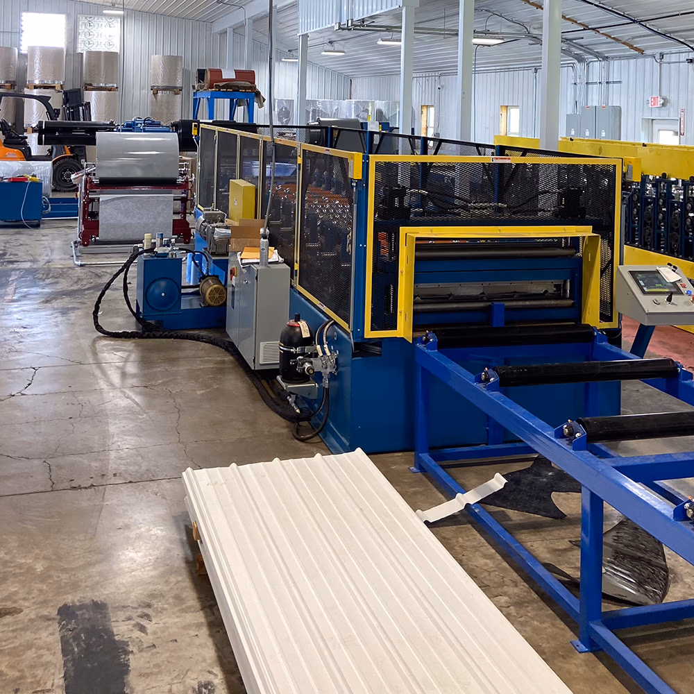 A blue and yellow roll forming machine in an industrial facility producing corrugated white metal roofing panels, with a steel coil decoiler visible in the background.