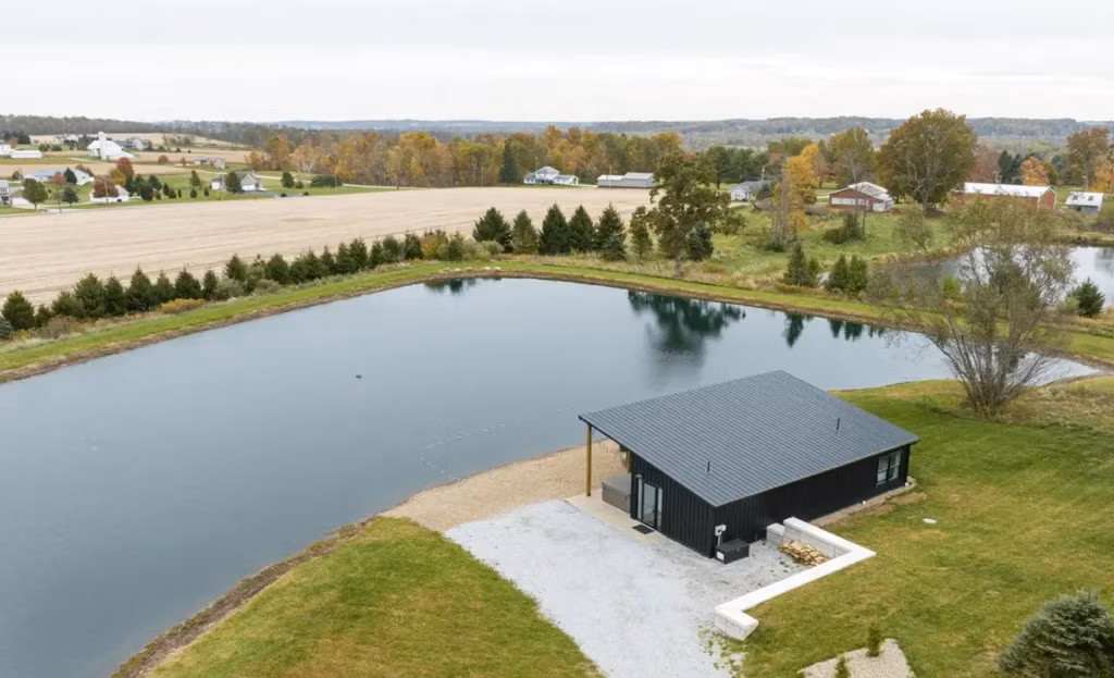 Aerial view of a modern black metal board and batten building with a gray standing seam roof situated beside a large private pond surrounded by green grass, gravel, and autumn trees in a rural Ohio landscape.