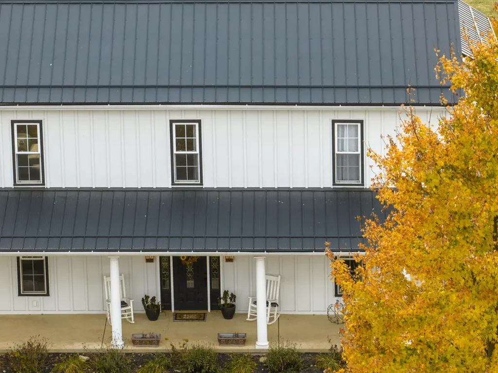 Aerial view of a white board-and-batten farmhouse with a dark charcoal standing-seam metal roof, a covered front porch with white columns and rocking chairs, and a yellow autumn tree in the foreground.
