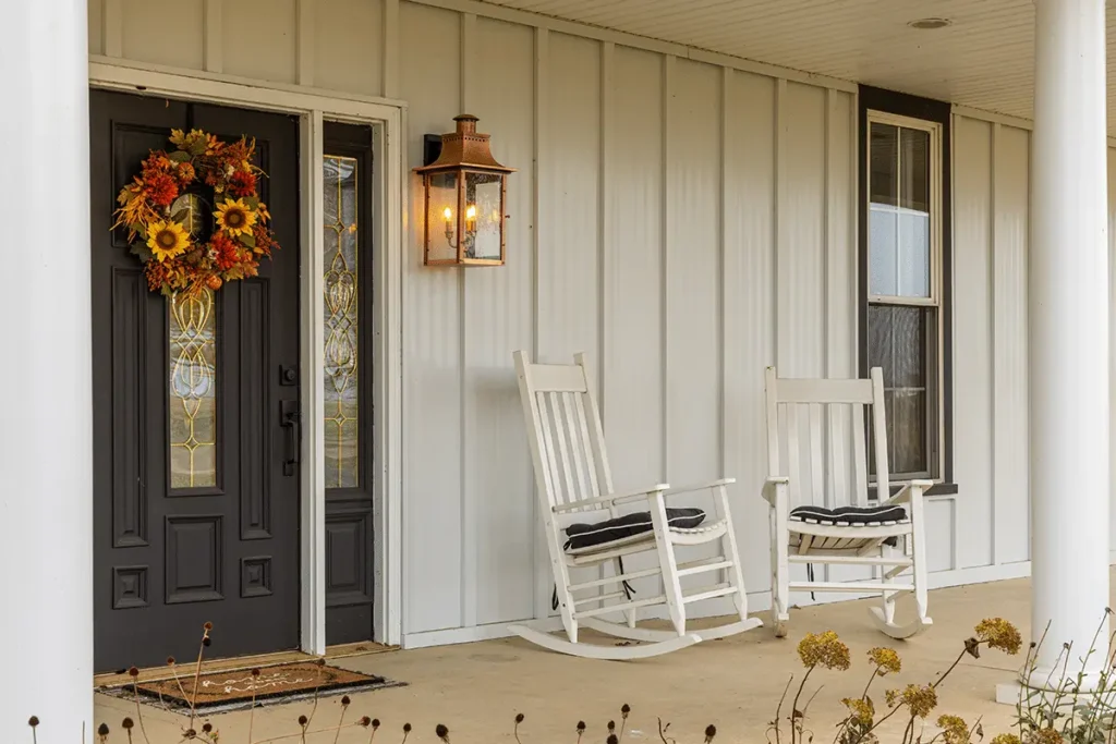 A white board and batten farmhouse exterior featuring a black front door adorned with a fall sunflower wreath, two white wooden rocking chairs with black cushions, and a lit copper lantern wall sconce on the covered front porch.