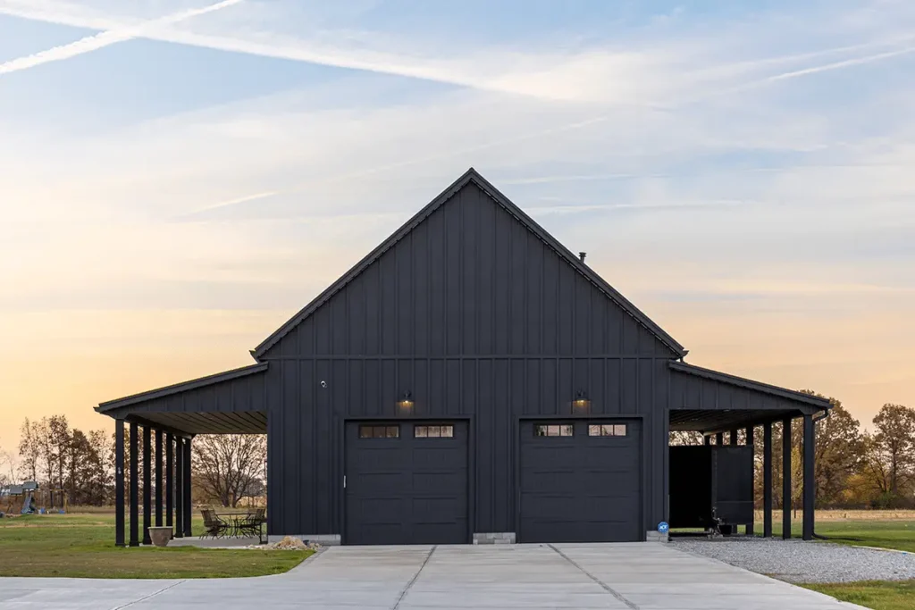 A striking all-black board and batten metal pole barn building featuring two matching garage doors with glass panels, flanked by open covered lean-to carports on each side, set on a concrete driveway at sunset in a rural Ohio landscape.