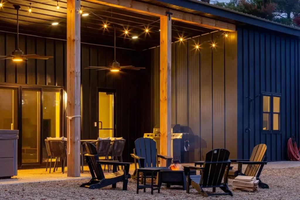 A modern navy blue corrugated metal barndominium exterior at dusk featuring a covered porch with string lights, ceiling fans, Adirondack chairs arranged around a glowing fire pit, and a gas grill on a gravel patio.
