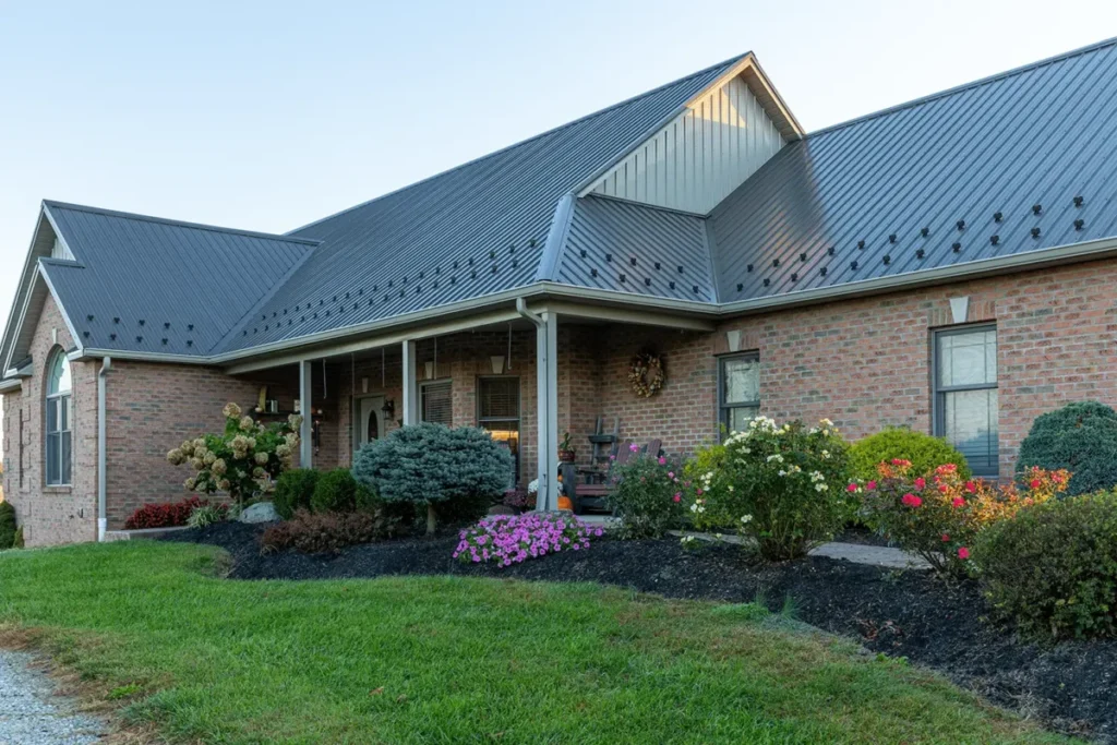 A brick ranch-style home with a dark gray standing-seam metal roof, covered front porch with rocking chairs, and lush landscaping featuring colorful flowers, trimmed shrubs, and black mulch beds.