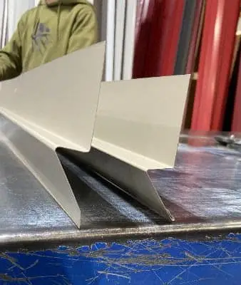 Two cream-colored metal rat guard trim pieces with angular bends stand upright on a steel fabrication table, with a worker in a green jacket visible in the background.