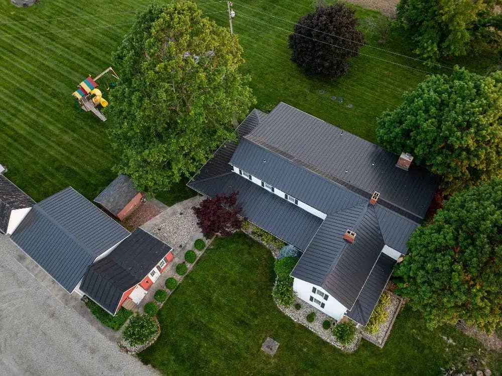 Drone aerial view of a white two-story home with dark charcoal standing seam metal roofing, surrounded by lush green lawn, mature trees, a red outbuilding, and a children's playset.