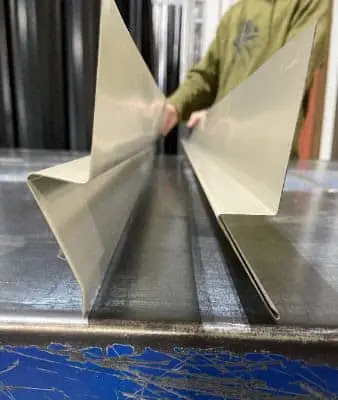 A person in an olive green shirt holds two freshly bent beige metal rat guard flashing pieces on a metal work table in a sheet metal fabrication shop.