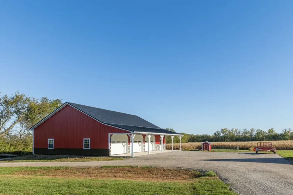 A large red metal pole barn with white trim, a covered open porch supported by white columns, and a dark metal roof, situated beside a mature cornfield under a clear blue sky on a rural farm property.