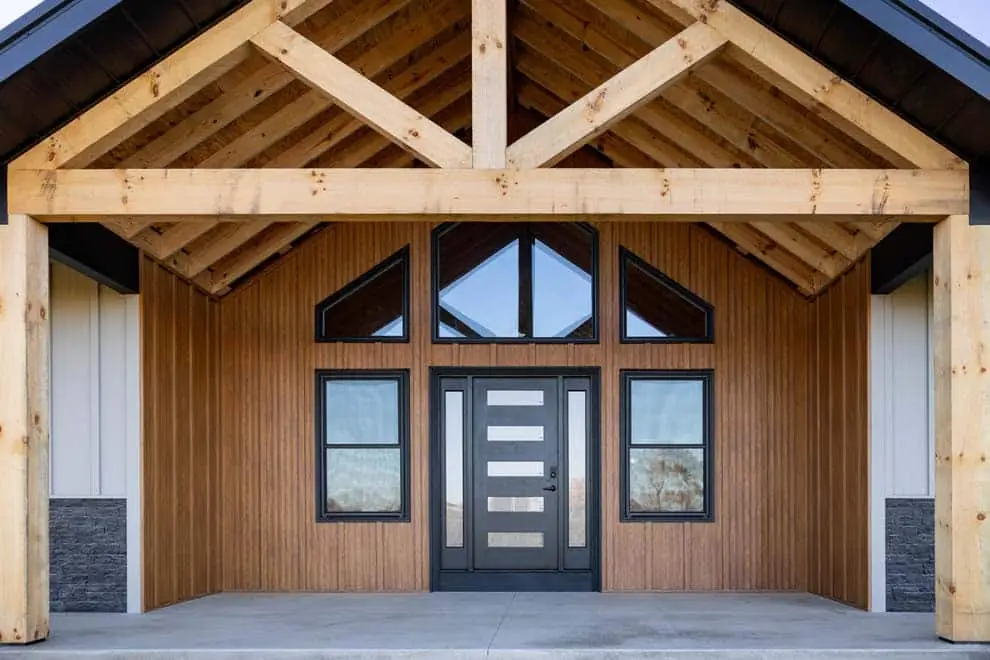A modern farmhouse entryway featuring warm brown wood grain corrugated steel board and batten siding, a black contemporary front door with horizontal glass panels, flanking windows, and an exposed timber frame gable porch with natural wood trusses and columns.