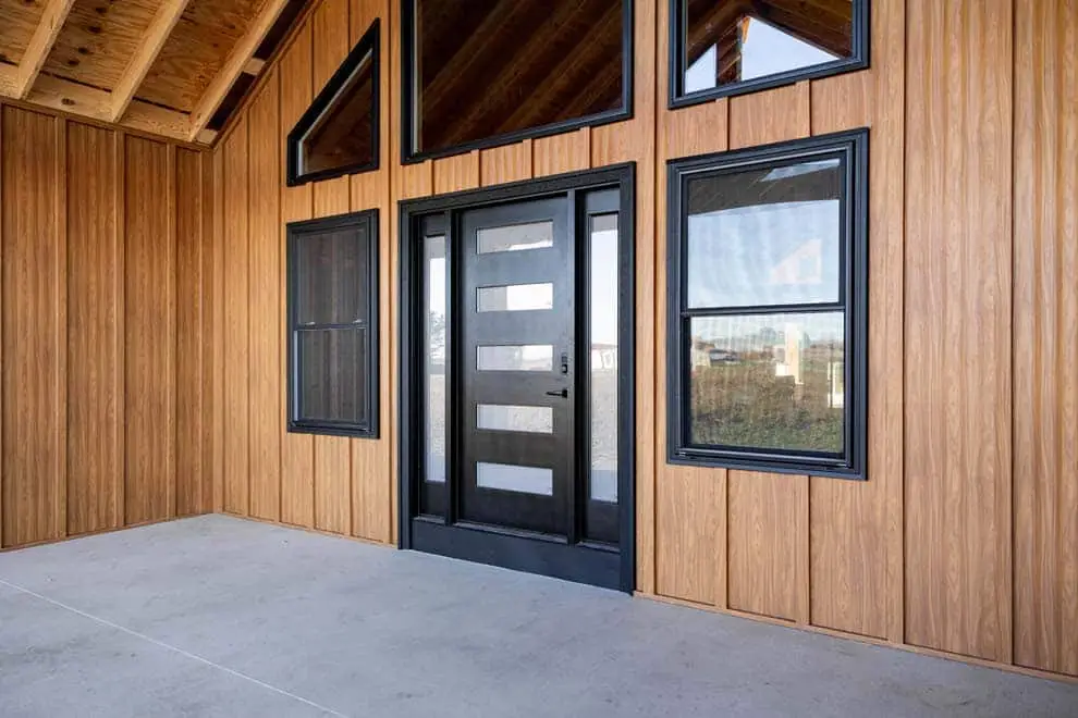 A modern home exterior featuring warm brown wood grain board and batten metal siding panels with a black contemporary front door with horizontal glass inserts, black-framed windows, and an exposed wood beam ceiling on a covered concrete porch.